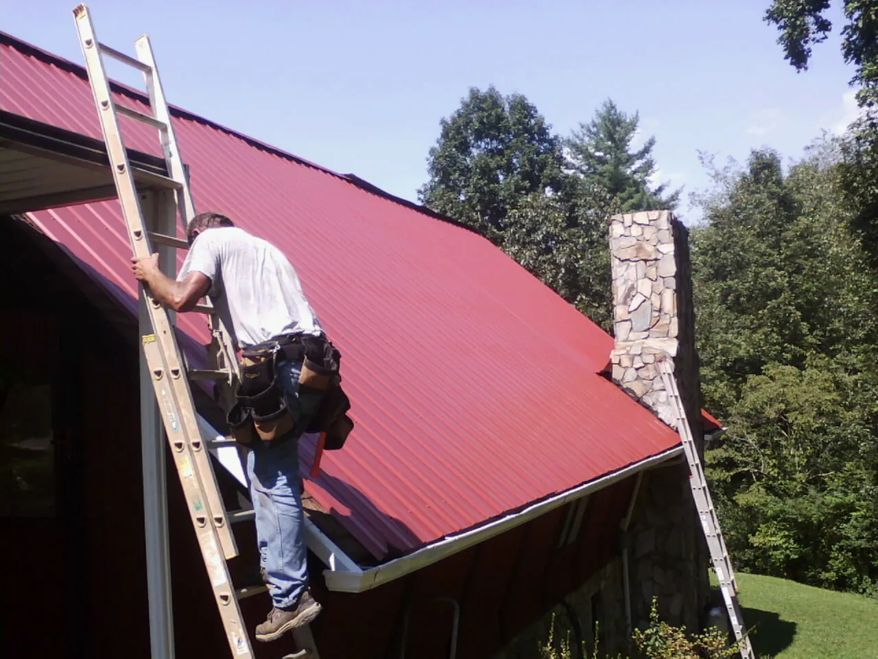Roofer on a ladder beside a red metal roof and stone chimney — Soffit & Fascia Repair by Roofing Brothers in New Holland